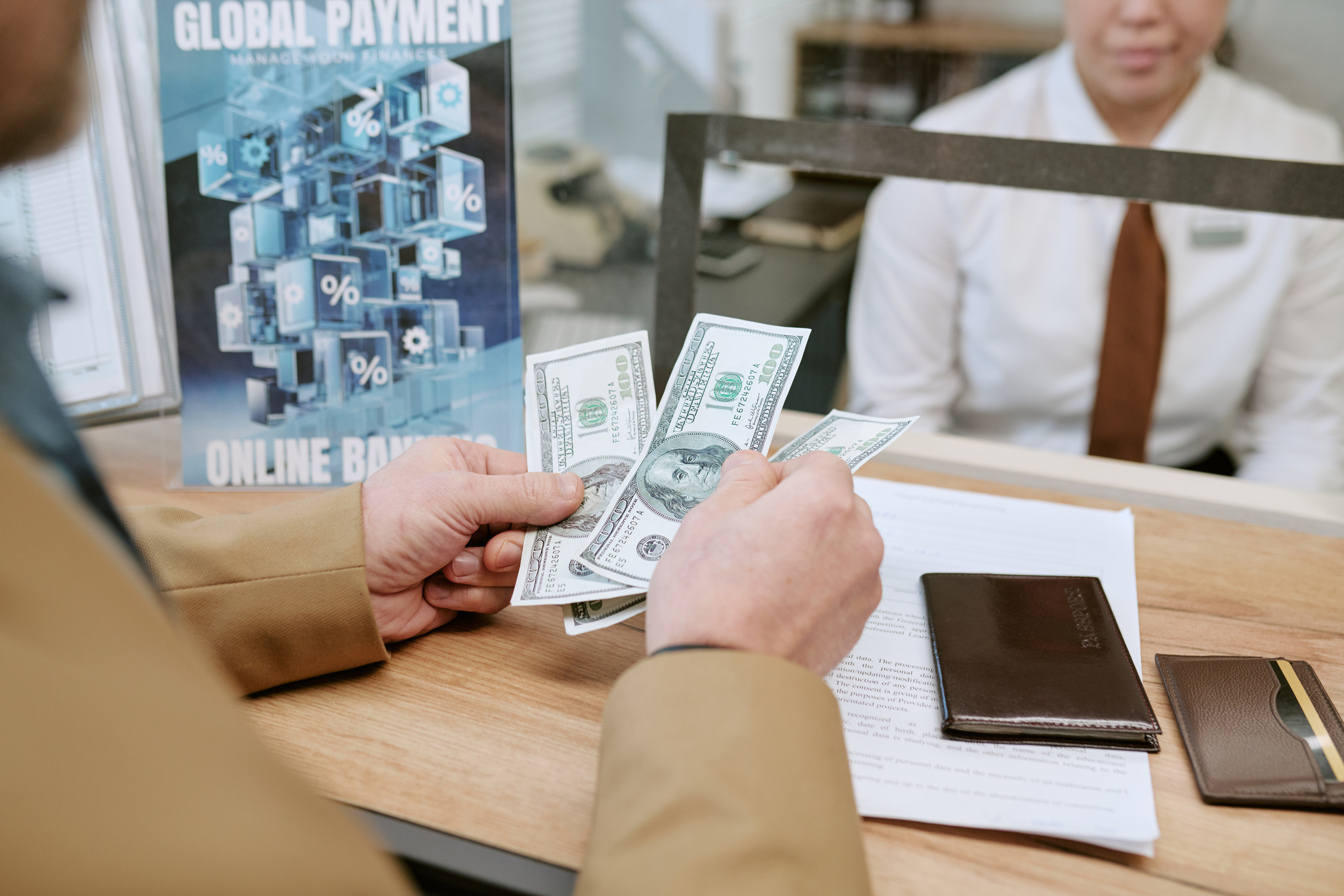 Middle aged Caucasian man counting US dollar bills at bank branch counter with female bank employee sitting behind glass partition, documents and wallet lying on wooden desk