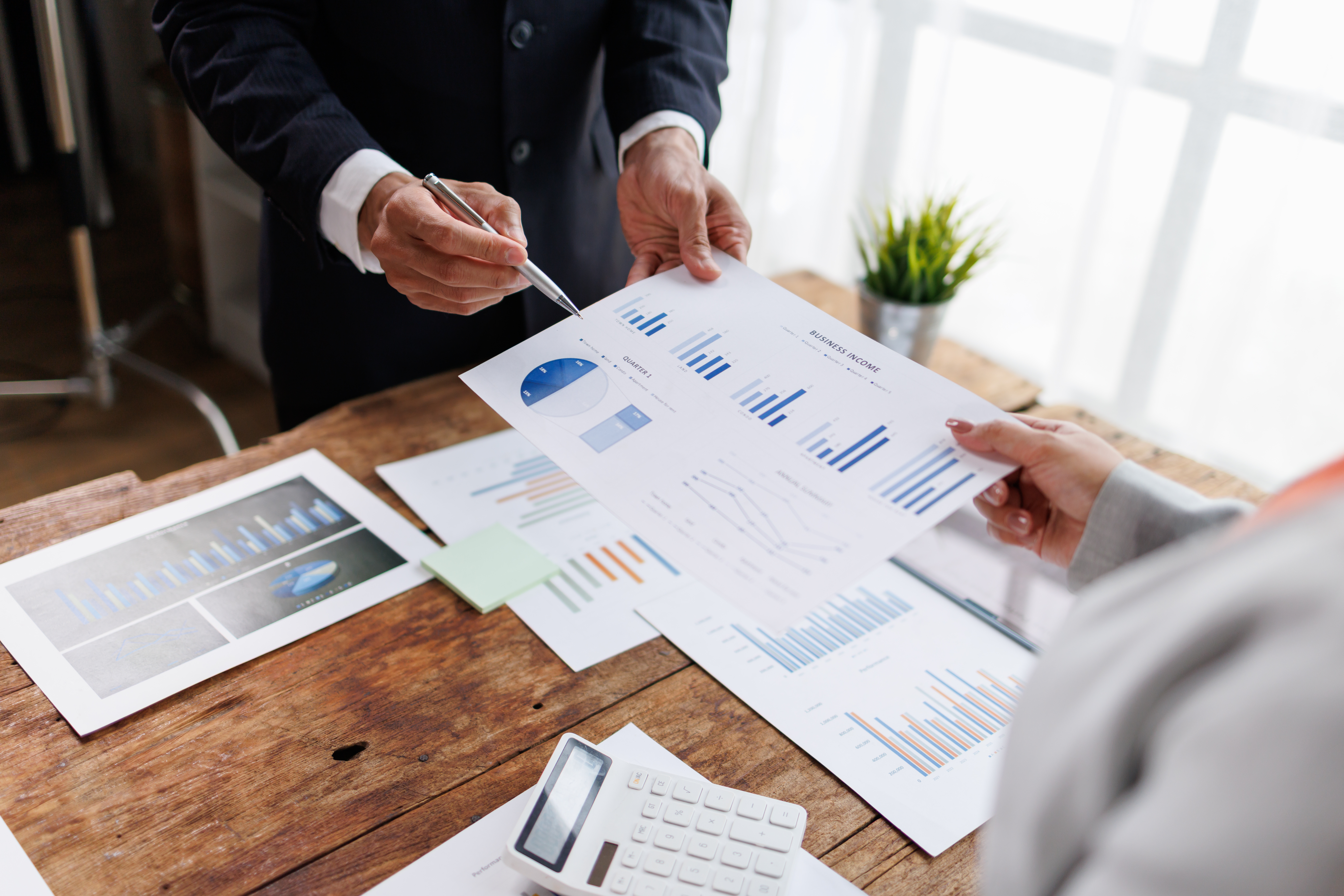 Business professionals are collaborating on financial reports. Analyzing data. And discussing investment strategies. Showing hands examining charts and graphs on a rustic wooden desk with a calculator